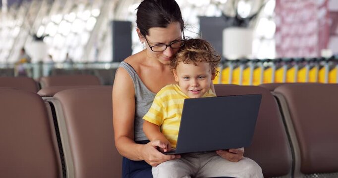 Mother with her little son sitting on her lap looks at photos on laptop while waiting for their flight in large airport. Toddler smiles with joy as he watches screen and presses buttons