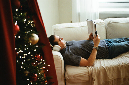 Man relaxing with book and headphones by Christmas tree