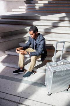Traveler checks phone while waiting on stairs with luggage.