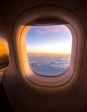 Aerial view of a sunrise from an aircraft window with clouds below. The sky transitions from orange to blue