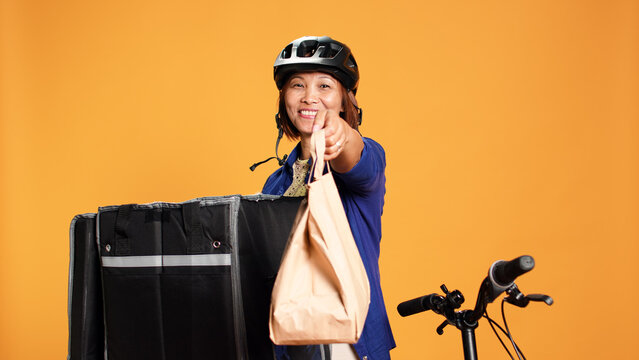 Portrait of asian woman taking lunch bag out of professional thermic backpack. Cheerful courier isolated over orange studio background handing customer food delivery order