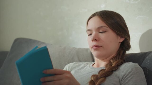 Woman reading blue book on couch with attentive expression, closeup sequences show concentration and page turning, braided hair and soft daylight create studious home atmosphere, portraying study