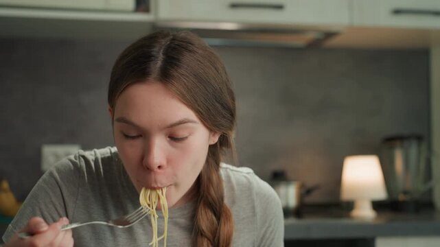 Pregnant caucasian woman eating noodles, at kitchen table, playful slurping, braided hair, casual tshirt, soft lamp light, spoon and plate visible, candid craving moment, cozy domestic atmosphere