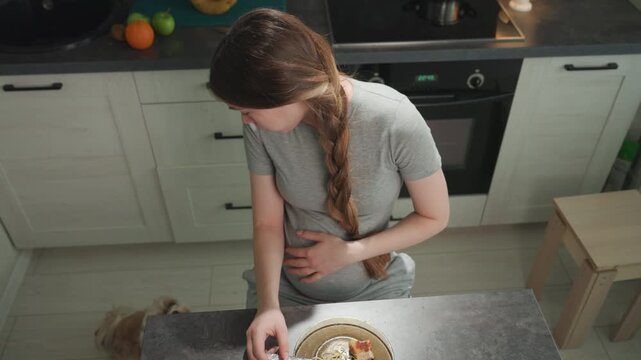 Pregnant caucasian woman in kitchen holds belly and reaches for bowl on counter, braided hair over shoulder, soft morning light, domestic calm, gentle craving and slight nausea while preparing small