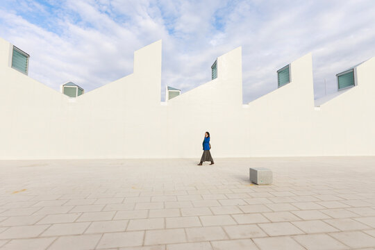 Person Walks in Open Space Near White Wall Under Cloudy Sky