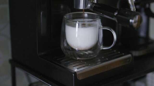 Modern coffee machine pouring a stream of hot espresso into frothed milk in a transparent glass cup, creating a layered cappuccino with rich crema and a distinct brown and white color separation