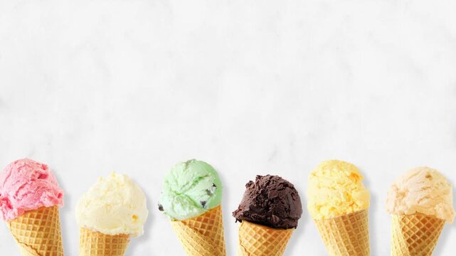 Ice cream cone moving bottom border with an assortment of flavors. Top down view, stop motion loopable video on a white marble background with copy space.