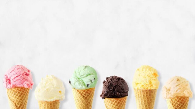 Ice cream cone moving bottom border with a selection of flavors. Above view, stop motion loopable video on a white marble background with copy space.