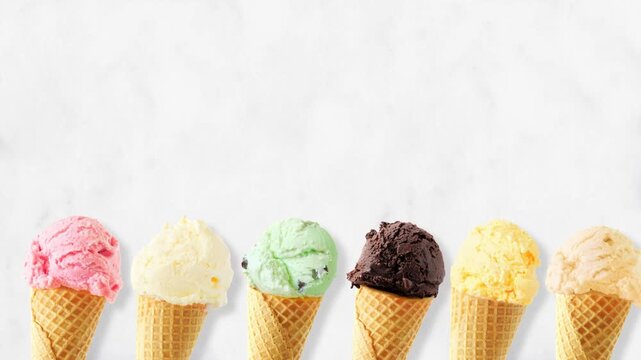 Ice cream cone moving bottom border with a variety of flavors. Overhead view, stop motion loopable video on a white marble background with copy space.