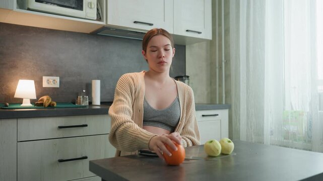 Pregnant caucasian woman at kitchen table holding orange and arranging apples, peeling banana, wearing cardigan and bralette, soft morning light through window, expectant mother planning healthy