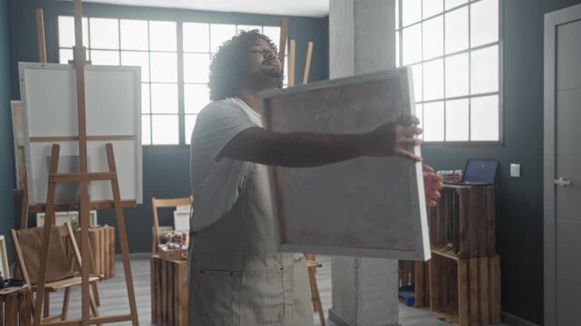 Man painter holds canvas with both hands in studio smiling while presenting an abstract painting; quiet pride creativity.