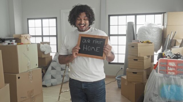 Young indian man smiling holds chalkboard reading our first home, hand on chest and looking aside amid stacked moving boxes, ladder and wrapped rug in a building; pride hope.