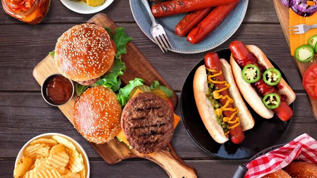 Summer BBQ food table scene with hot dog and hamburger buffet. Stop motion loopable table placement video. Top view over a dark wood background.