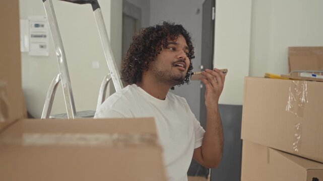 Young indian man holds smartphone to ear, smiling and talking beside stacked cardboard boxes and an aluminum ladder in a new building; moving day joy.