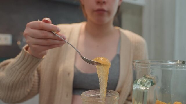 Pregnant woman scooping golden honey spoon. Closeup focus on sticky drizzle and viscous texture, jar and glass on countertop, soft cardigan, intimate sensory moment, natural remedy and prenatal taste