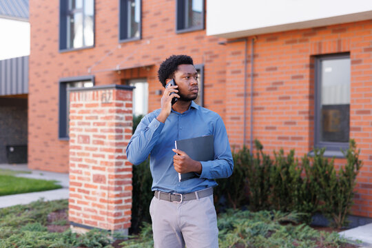Focused black male realtor talking on mobile phone near townhouse