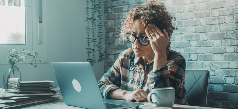 Stressed woman working on laptop in a home office surrounded by paperwork and coffee