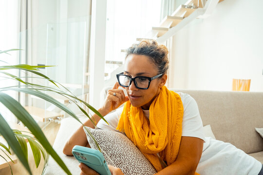 Woman with glasses relaxing on couch at home reading her phone while wearing a yellow scarf