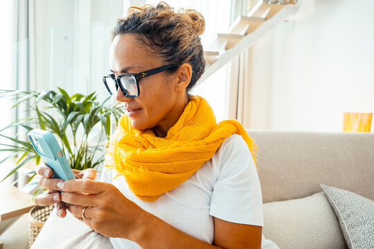 Woman in yellow scarf sitting on couch and using smartphone while relaxing at home