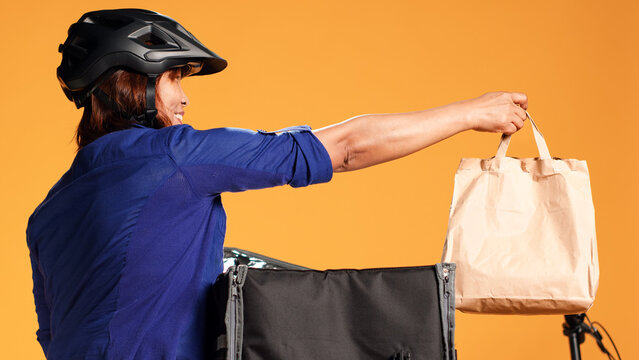 Close up shot of asian courier offering customer lunch bag order. Food delivery friendly worker arriving quick to client address, taking meal out of thermic backpack, isolated over studio background