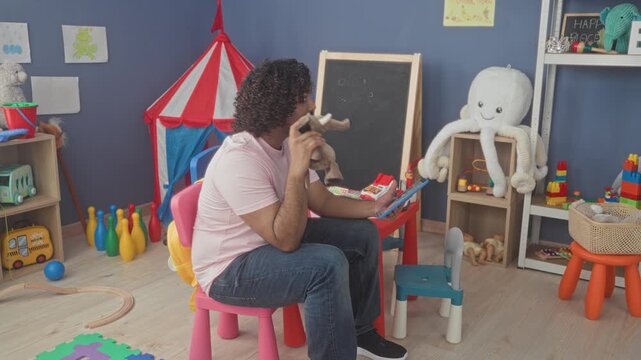 Man holds tablet and stuffed elephant puppet while seated in a kindergarten playroom inside a school building; playful care.