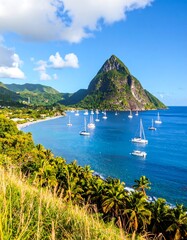 Obraz premium A vibrant coastal scene features a steep, green mountain rising from the azure ocean. Numerous sailboats dot the turquoise water