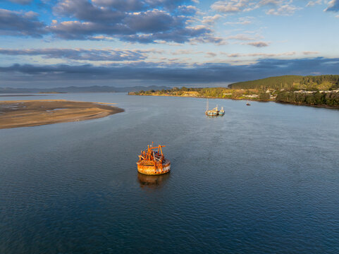 A large, rusty dredging barge sits in the calm waters of the Kawhia Harbour, Kawhia, Waikato, New Zealand, at sunrise. The barge is used for seabed maintenance and resource extraction.