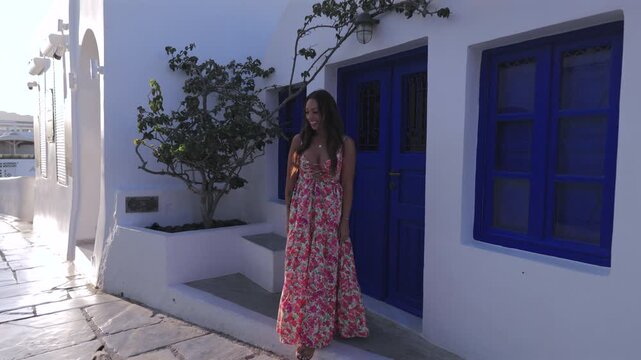 Beautiful young woman wearing a colorful floral maxi dress stands smiling in front of traditional white Greek architecture with vibrant blue doors and windows. A cat walks by on the stone street.