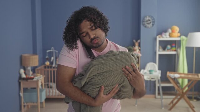 Man holds phone to ear and cradles wrapped baby in blanket with his hand in studio; parenting exhaustion.