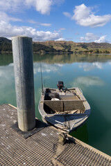 Fototapeta premium A small fishing boat with an outboard motor is moored to a jetty on a calm, reflective bay in Kawhia Harbour, Te Waitere, Waikato, New Zealand. The boat is ready for a day of fishing.