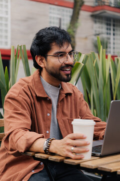 Young man using laptop and holding coffee at outdoor cafe