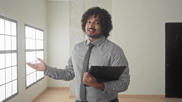 Young man holds folder and gestures palm up while presenting an empty house building for sale; friendly trust.