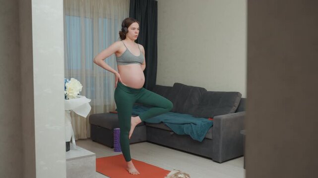 Pregnant white woman standing on mat in cozy living room, hands on hips, balancing tree pose, soft morning light, couch and flowers in background, focused calm breathing for prenatal fitness