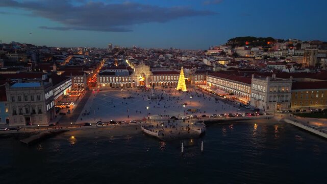 Drone perspective shows illuminated central plaza in Lisbon, tall festive tree, surrounding historic facades and calm river reflections under deep blue evening sky creating strong holiday atmosphere