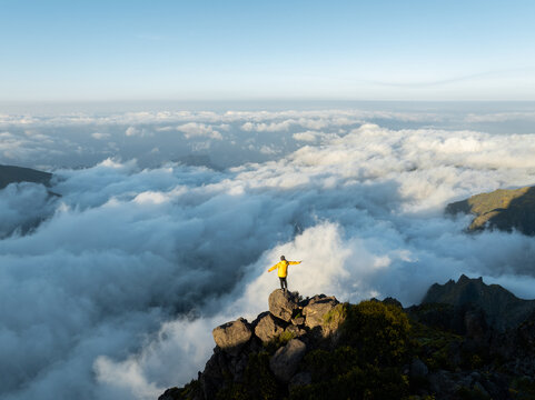 Aerial view of a person in a yellow jacket standing on the rocky summit of Pico do Arieiro above a thick sea of clouds and rugged mountain peaks in Curral das Freiras, Madeira, Portugal.
