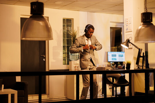 Cheerful worker wearing headphones showing dance moves in startup office during work break. Positive entrepreneur dacing and having fun after finishing company strategy, planning investment plan