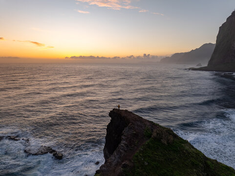 Aerial view of a person standing on the edge of Miradouro do Guindaste cliff overlooking the Atlantic Ocean during a golden sunrise in Faial, Madeira, Portugal.