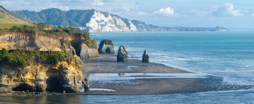Two people walk along the beach at the The Three Sisters, Tongaporutu, New Zealand, with its iconic rock formations and sea stacks. The sun illuminates the layered cliffs.