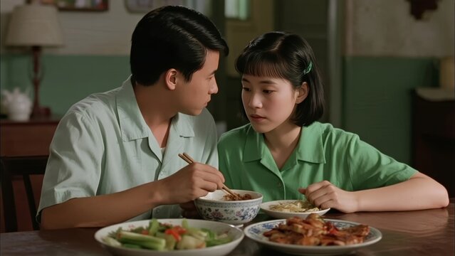 A young couple shares a meal at a wooden dining table, eating with chopsticks in a modestly furnished room.