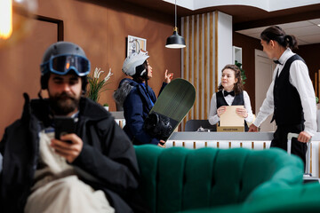 Naklejka premium At front desk, employees helping young woman with snowboard equipment with her room reservation at winter resort. Man with ski helmet on sofa uses his mobile phone while female tourist does check-in.