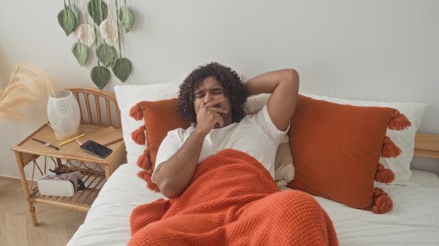 Man lying on bed with orange blanket clutching chest and hand on throat beside bedside table and smartphone in studio; discomfort rest.