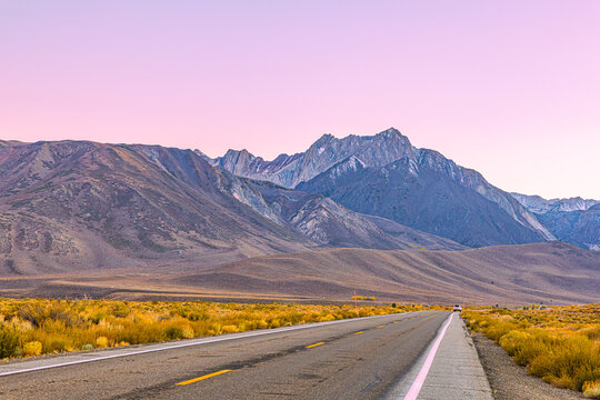 A lonely two-lane highway stretches toward the rugged Eastern Sierra Nevada peaks under a soft pink twilight sky near Mammoth Lakes, California.