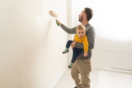 Father holding son painting home wall with roller