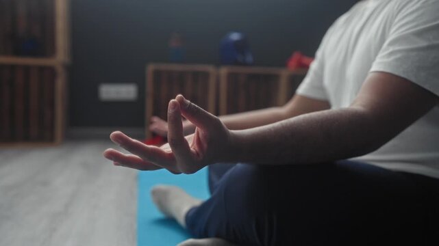 Man seated crosslegged meditating with hand in gyan mudra on blue yogamat at studio near wooden shelf with helmet and gloves visible; serenity focus.
