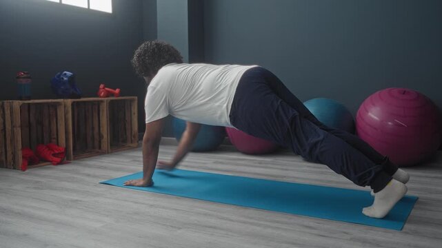 Man doing plank with shoulder tap on blue yoga mat in studio by window with exercise balls, wooden crate, red dumbbell and bottle; focus strength.