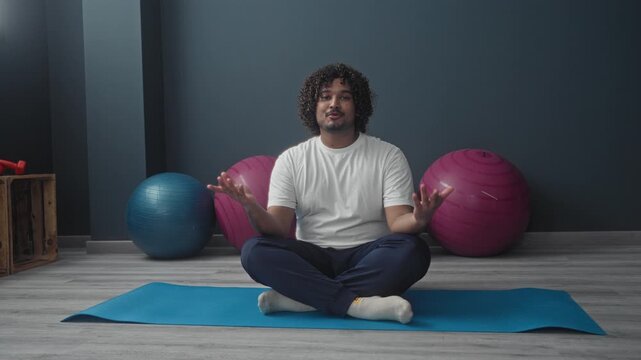 Man seated cross legged on a blue yoga mat raises both hands palms open beside exercise balls in a building; mindful calm.