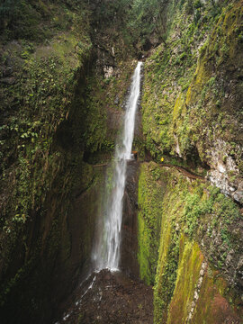Aerial view of a tall waterfall cascading down lush mossy cliffs with a person in a yellow jacket on a hiking trail in Madeira, Portugal.