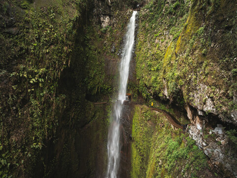 Aerial view of a tall waterfall cascading down a lush green canyon with a person walking on a narrow levada path Madeira, Portugal.