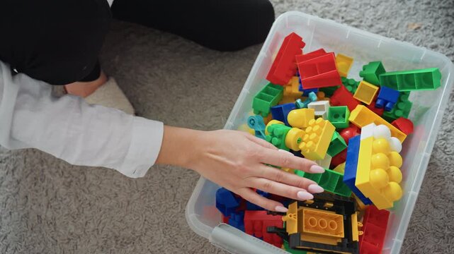Mom kneeling by toy bin sorting colorful blocks and small bricks, closeup of hands reaching into plastic container on carpet, gentle tidy routine after play, soft slippers and bedroom corner visible,
