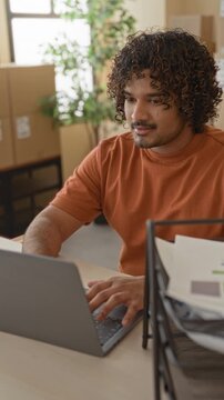 Man typing on laptop and holding pen beside stacked cardboard boxes and a barcode scanner while arranging packages in a building; small business focus.